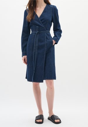 Femme portant une robe portefeuille en denim bleu foncé à manches longues et longueur genou, avec ceinture, poches latérales et sandales noires à enfiler, debout sur fond blanc.