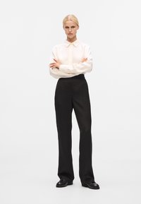 Camicia bianca a maniche lunghe con colletto a punta, abbinata a pantaloni neri a vita alta. I pantaloni presentano un motivo tessurizzato e un taglio dritto.