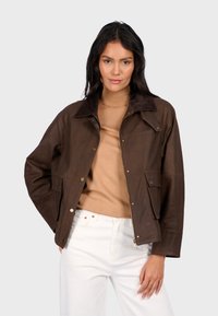 Hnědá kožená bunda s límcem z kožešiny, se dvěma předními kapsami a zlatými patentkami. Následující je nošena přes béžový svetr a bílé džíny.