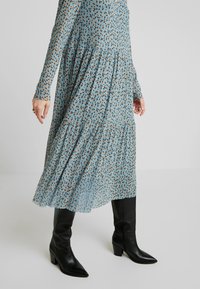 Niebieska sukienka w wzory z długimi rękawami, z warstwowym krojem i małymi kwiatowymi detalami w kolorze pomarańczowym i brązowym; zestawiona z czarnymi botkami na obcasie.