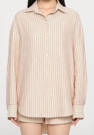 Personne portant une chemise beige ample à manches longues, boutonnée, avec de fines rayures verticales marron et un short assorti.