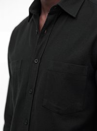 Gros plan d'une personne portant une chemise noire à boutons avec col et poche poitrine sur un fond blanc.