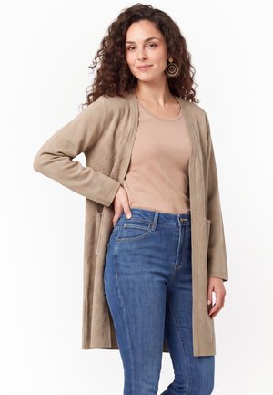Kvinde med krøllet hår iført beige lang cardigan, tan top, blå jeans og store runde øreringe, smilende let med hånden på hoften.