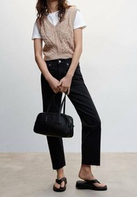 Femme portant un gilet en tricot beige sur un t-shirt blanc, un jean noir taille haute, des sandales compensées noires, tenant un petit sac à main noir.