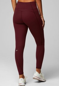 Weinrote, hoch taillierte Leggings mit einer glatten Textur und Flachnähnen, versehen mit einem kleinen Logo nahe dem Knöchel und kombiniert mit weißen Turnschuhen.