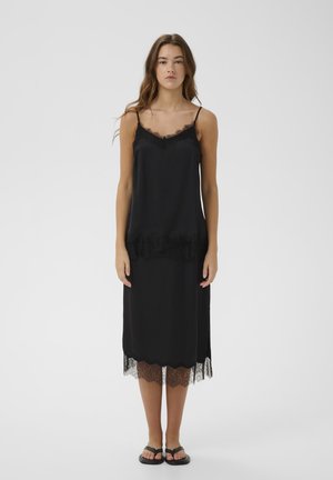 Kobieta ubrana w czarną sukienkę na ramiączkach z koronkową lamówką przy dekolcie i na dole, stojąca na tle jednolitego białego tła.