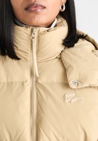 Giacca imbottita beige con colletto alto, chiusura con zip e bottoni a pressione. Presenta un logo ricamato sul fronte e un tessuto strutturato.