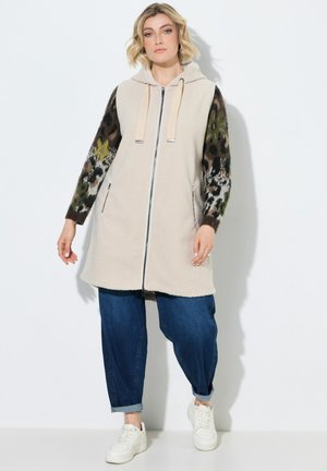 Donna che indossa un gilet beige senza maniche con cerniera e cappuccio sopra una maglia a maniche lunghe con fantasia, jeans blu con risvolti e sneakers bianche, in piedi su sfondo bianco.
