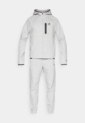 Conjunto deportivo gris claro que incluye sudadera y pantalones, con cierre frontal con cremallera, capucha y detalles negros en los puños y el bolsillo del pecho. Textura suave.