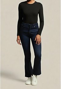 Personne portant un haut noir à manches longues, un jean flare taille haute bleu foncé et des baskets blanches, debout devant un fond uni.