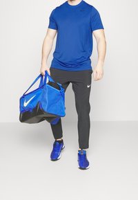 Modré sportovní tričko s krátkým rukávem, černé kalhoty a modré tenisky. Nese velkou modrou tašku přes rameno s černými akcenty a logem.