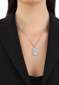 Collana con pendente in argento di forma ovale, decorata con intricate embellishment simili a diamanti, attaccata a una delicata catena, indossata su un outfit nero.