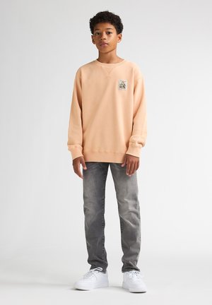 Sweatshirt pêche clair à col rond, avec une petite écusson imprimé sur la poitrine gauche, associé à un jean gris et des baskets blanches.