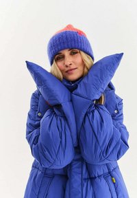 Modrá prošívaná bunda s nadměrně velkými rukávy, detaily s knoflíky a vysokým límcem. Pletená čepice je fialová a růžová.