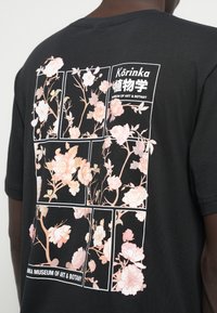 Μαύρο μπλουζάκι με floral σχέδιο σε ροζ και λευκές αποχρώσεις, με ορθογώνια γραφική απεικόνιση που περιλαμβάνει κείμενο σχετικά με ένα μουσείο.