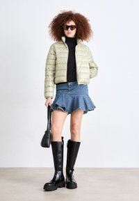 Polstret olivengrønn jakke, svart turtleneck, denim flare-skjørt, svarte høye støvler og svart håndveske. Modellen har krøllete hår og solbriller.