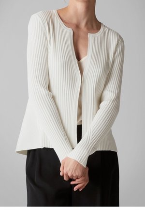 Person iført en hvid ribbet cardigan over en lys top med sorte bukser, stående med hænderne foldet foran mod en ensfarvet baggrund.