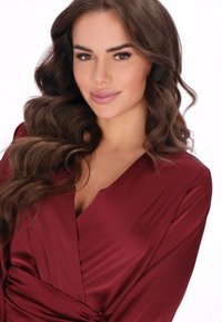 Hedvábná bordó šaty s výstřihem do V, zavinovacím designem a splývavými rukávy. Hladká textura s jemným leskem zvýrazňuje střih.