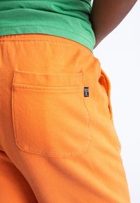 Oranje katoenen broek met een zichtbare achterzak, voorzien van een klein zwart merkje. De tailleband is elastisch voor extra comfort.