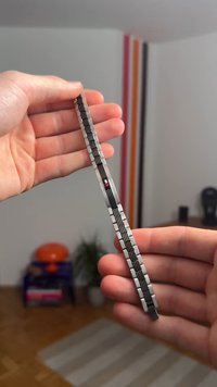 Metāla lineāls ar elegantu sudraba apdari, melniem atzīmējumiem, izcelta tekstūra un neliela sarkana aplīša detaļa. Turēts rokā.