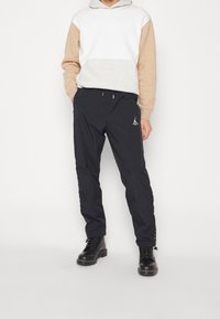 Felpa grigia e beige con tasca a marsupio, abbinata a pantaloni jogger neri con logo ricamato. Entrambi gli articoli hanno una vestibilità comoda.