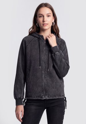 Jonge vrouw met lang haar, een zwarte zip-up hoodie met trekkoorden en zwarte broek, staand tegen een eenvoudige lichte achtergrond.