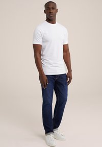 Camiseta blanca de manga corta, corte ajustado, combinada con jeans oscuros y zapatillas blancas, sobre un fondo claro.
