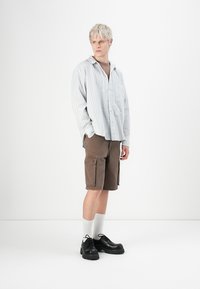 Young person in oversized white striped shirt, brown cargo shorts, white socks, and black lace-up shoes standing against a white background.