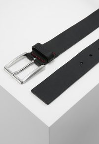 Cintura in pelle nera con fibbia metallica argento. Presenta cuciture rosse lungo il bordo e fori regolabili lungo la cintura.