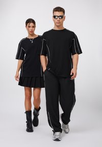 Juodos didelės t-shirts su baltomis siuvinėjimo detalėmis, derinamos su juoda raukšlėta sijonų ir kelnių aukščio batais. Vyrų modelis vilki laisvas juodas kelnes ir sportinius batus.