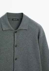 Grijze gebreide cardigan met een kraag en knoopsluiting. Voorzien van zwarte knopen, textiel met structuur, en een label bij de kraag.