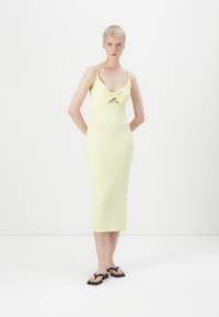 Robe midi jaune avec des fines bretelles, détail découpé au niveau de la poitrine, coupe ajustée et texture lisse, associée à des sandales noires.