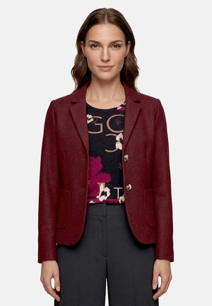 Femme portant un blazer bordeaux par-dessus une chemise noire à motifs floraux et textuels et un pantalon gris foncé, debout devant un fond uni.