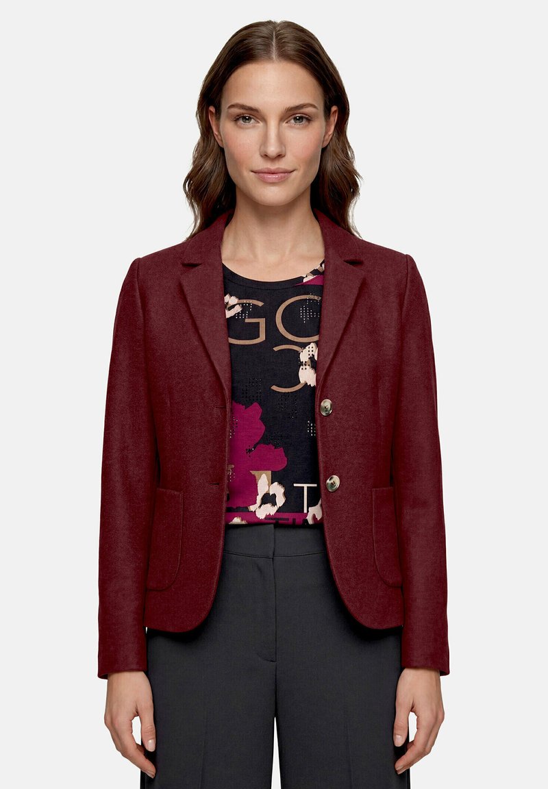 Femme portant un blazer bordeaux par-dessus une chemise noire à motifs floraux et textuels et un pantalon gris foncé, debout devant un fond uni.