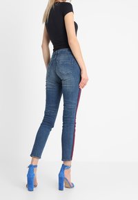 Jeans ajustados en denim azul oscuro con rayas laterales rojas, con una cintura de altura media y dos bolsillos traseros. Combinados con zapatos de tacón altos azules.