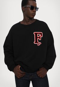 Zwarte sweatshirt met een oversized fit en ronde hals, voorzien van een rode geborduurde "F"-logo en een lippenafbeelding op de linkerkant van de borst.