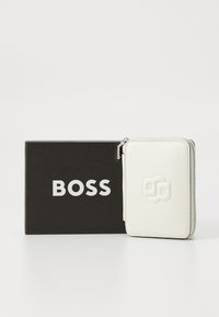 White leather wallet with embossed logo, zip closure, and textured surface, displayed beside a black box featuring white lettering.