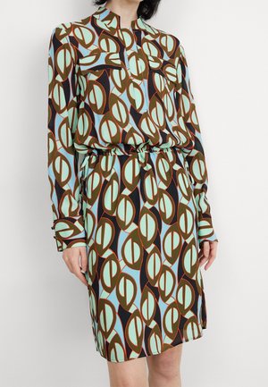 Vrouw die een lange mouwen jurk draagt met een geometrisch patroon in groen, lichtblauw, zwart en bruin, met een ceintuur in de taille en voorzakken.