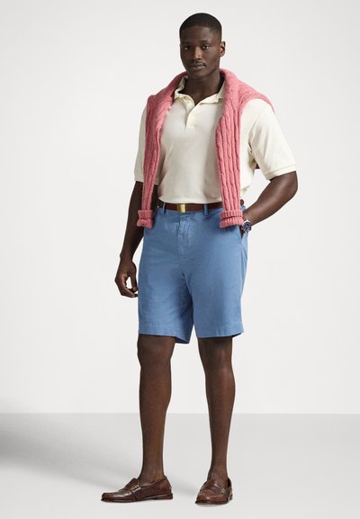 Gaiši bēša polos kreklu pāri zilām šortam. Pār pleciem uzmests rozā adīts džemperis. Brūnas mokasīnu kurpes ar izteiktām šuvēm.