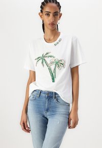 T-shirt bianco in cotone con design di palme in paillettes verdi e argento, abbinato a jeans in denim chiaro e distressed.