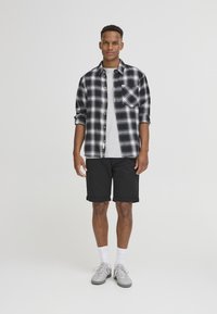 Svart og hvit rutete flanellskjorte over en grå t-skjorte, parret med svarte shorts og grå joggesko med hvite sokker. Enkelt design og avslappet passform.