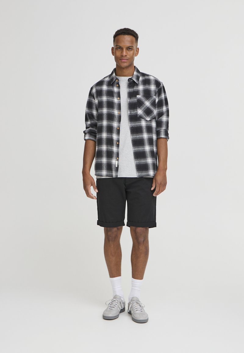 Svart og hvit rutete flanellskjorte over en grå t-skjorte, parret med svarte shorts og grå joggesko med hvite sokker. Enkelt design og avslappet passform.