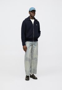 Marinblå stickad zip-up-tröja med vit krage, ljusblå keps, ljusa denimbyxor och bruna skor med blank finish.