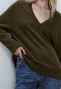 Olijfgroen V-halstrui van zacht materiaal. Heeft een relaxte pasvorm, geribbelde manchetten en een iets langere achterzoom. Gecombineerd met blauwe spijkerbroeken.