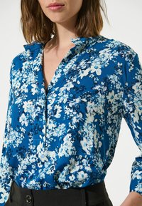 Bluse mit floralem Muster, die einen blauen Hintergrund, weiße und marinefarbene Blumen, einen Kragen und eine Knopfleiste aufweist, aus glattem Stoff gefertigt.