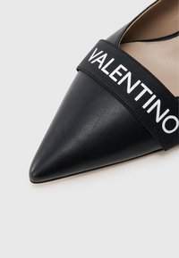 Musta nahkainen kärjestä terävä kenkä, jossa on teksturoitu päällinen ja leveä joustava nauha, jossa valkoisella kohokuvioitu "VALENTINO".