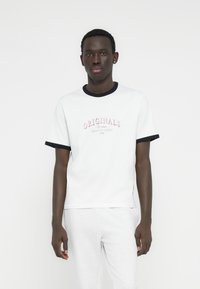 Λευκό βαμβακερό μπλουζάκι με σκούρο μπλε γιακά και περιθώριο στα μανίκια. Διαθέτει ροζ σχέδιο με τη λέξη «ORIGINALS» μπροστά. Απλός σχεδιασμός με στρογγυλή λαιμόκοψη.