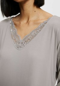 Top gris claro con escote en V que presenta un delicado borde de encaje gris. La tela es suave, con mangas largas, resaltando un ajuste relajado.
