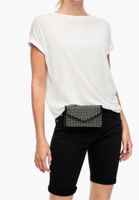 Borsa a tracolla in finta pelle nera con borchie argentate, chiusura a patta ripiegata. Il modello indossa una maglietta bianca e pantaloni corti neri arrotolati. Sfondo semplice.