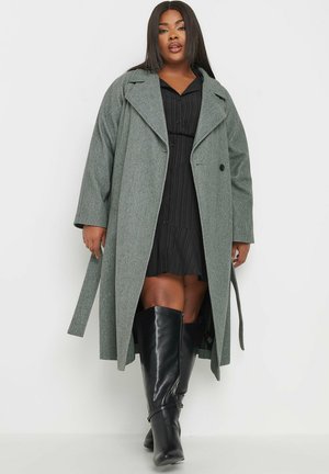 Grauer Wollmischung langer Mantel mit breiten Revers, einem Knopfverschluss und einem Bindegurt, kombiniert mit einem schwarzen, plissierten Kleid und kniehohen Stiefeln.